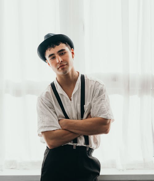 A man in suspenders and a hat is standing by a window