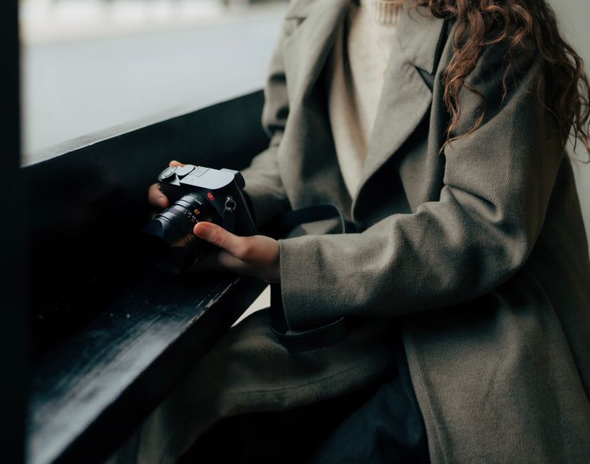 Elegant woman in coat holding camera indoors, showcasing stylish and vintage look.