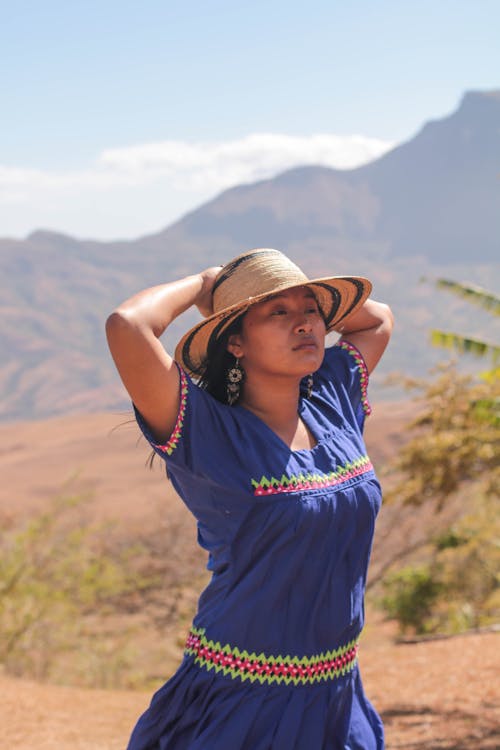 Young Woman in a Dress and Hat Standing in Sunlight on the Background of Mountains 