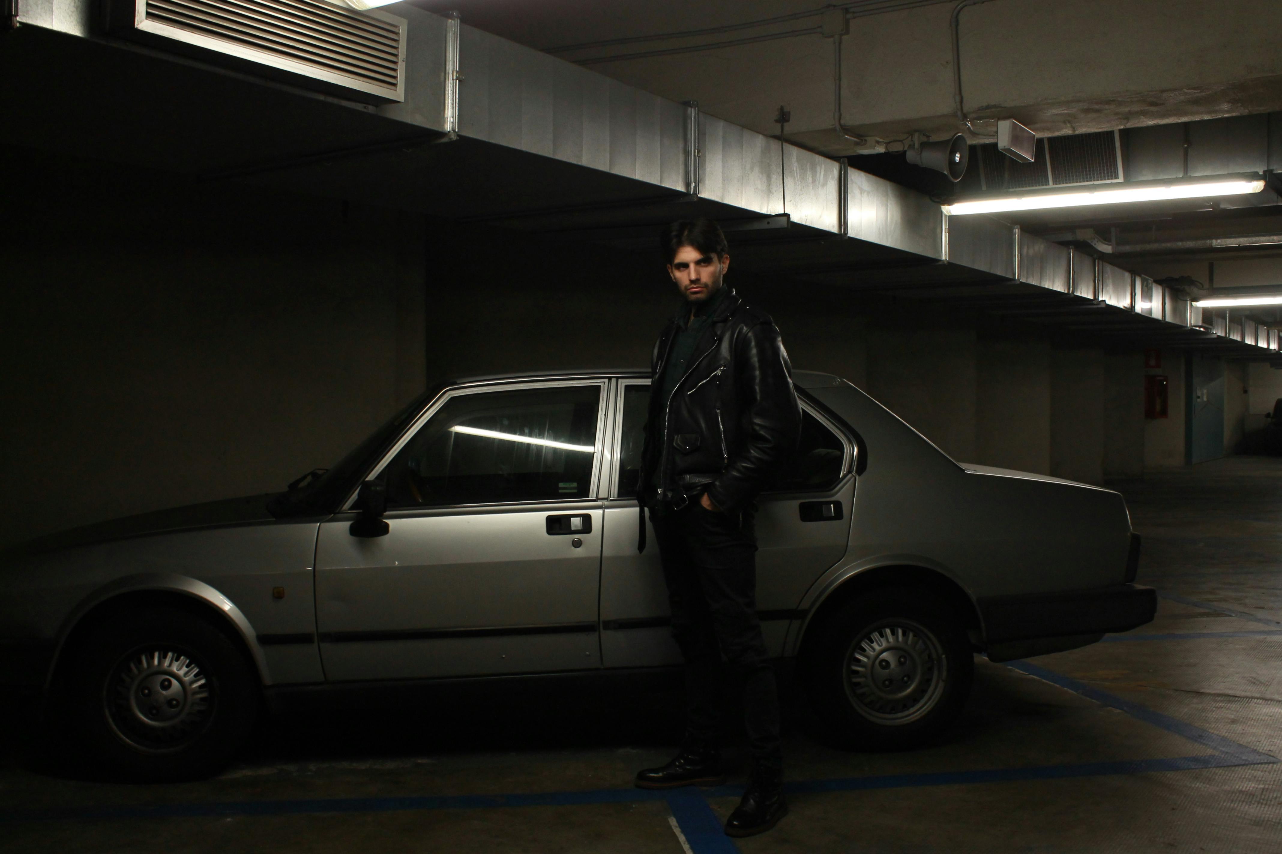 Man in leather jacket stands beside vintage car in dimly lit parking garage.