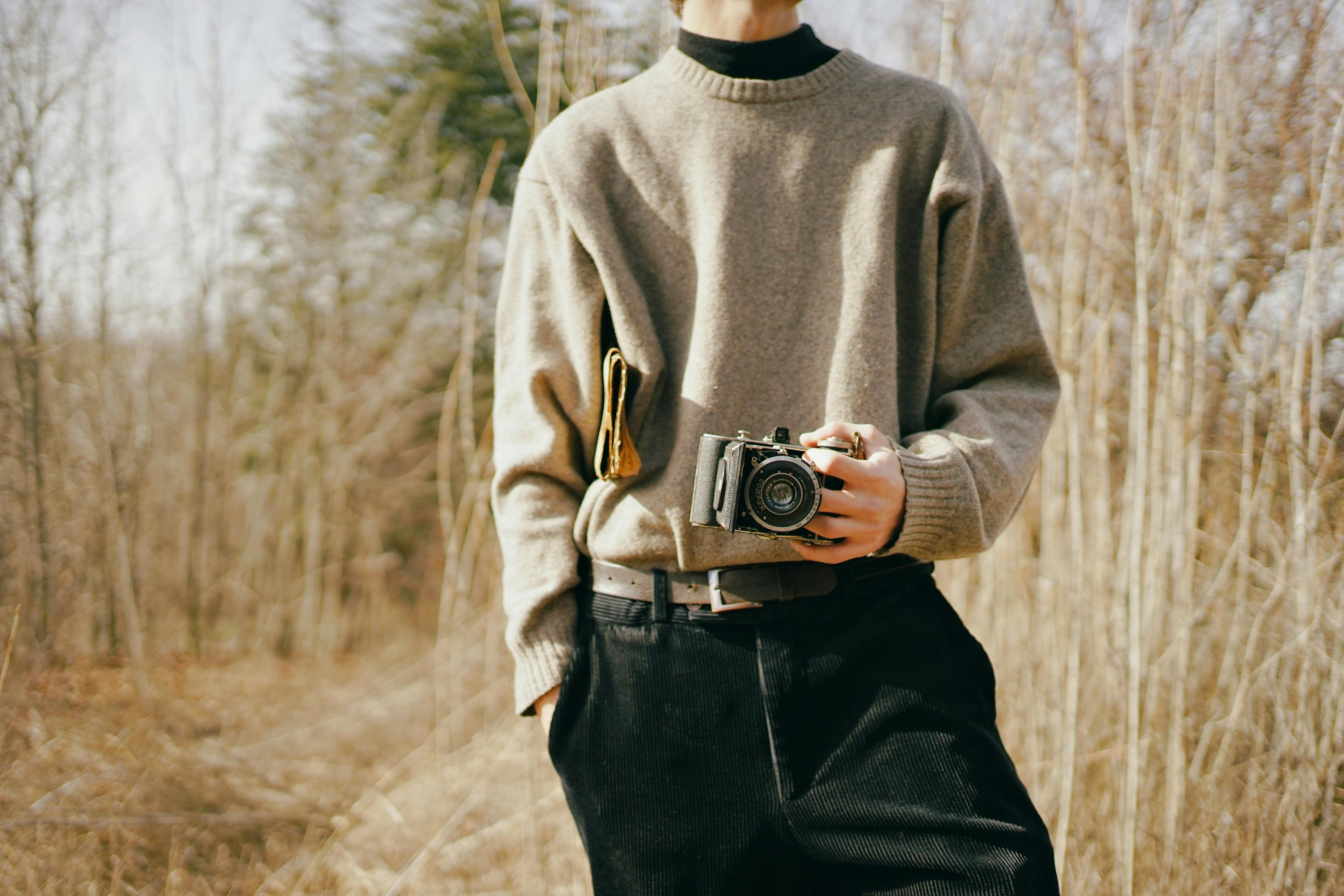 A man in a gray sweater holds a vintage camera in a rural Ankara landscape.