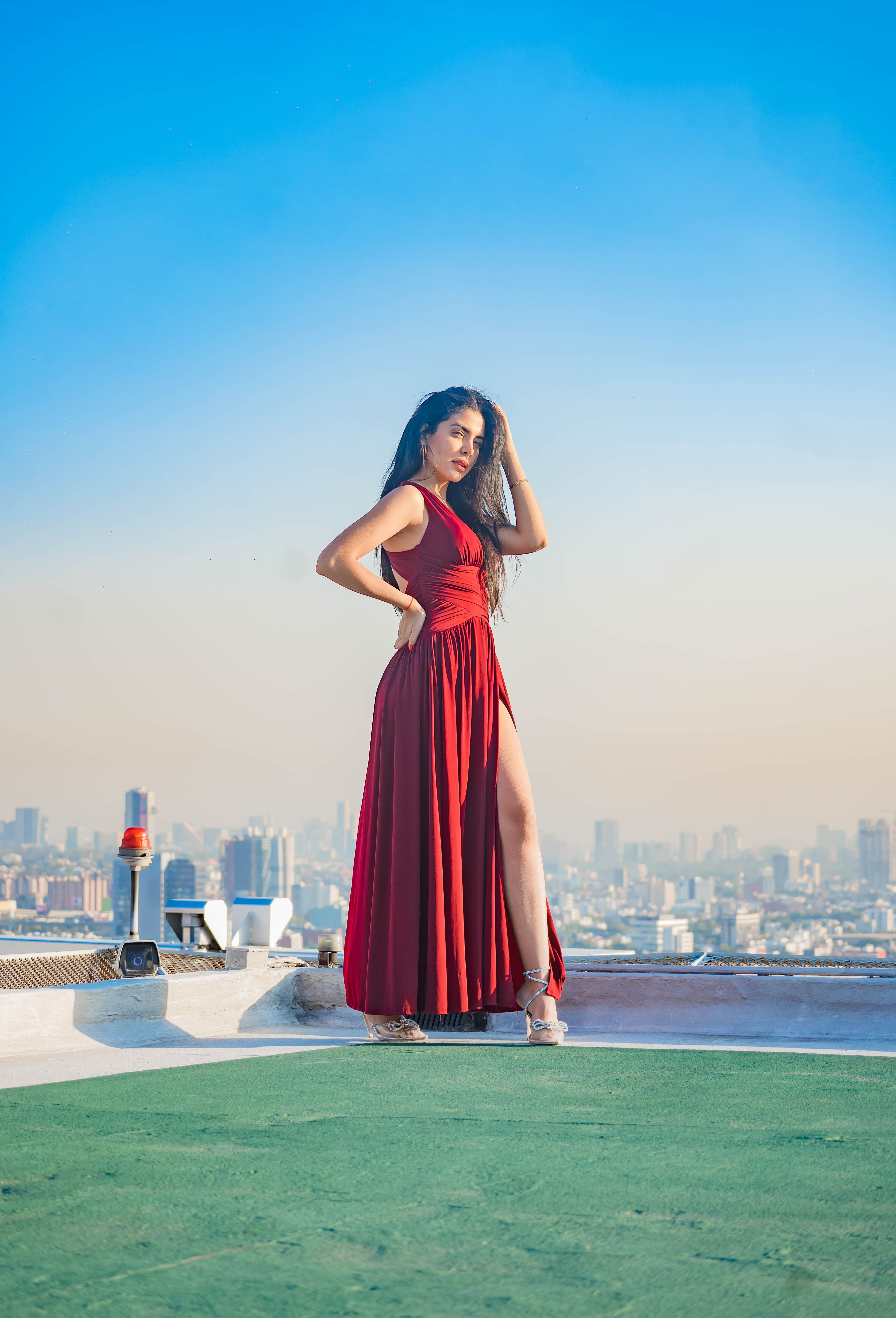 Elegant woman in a red dress posing confidently on a rooftop with a clear sky and city skyline in the background.