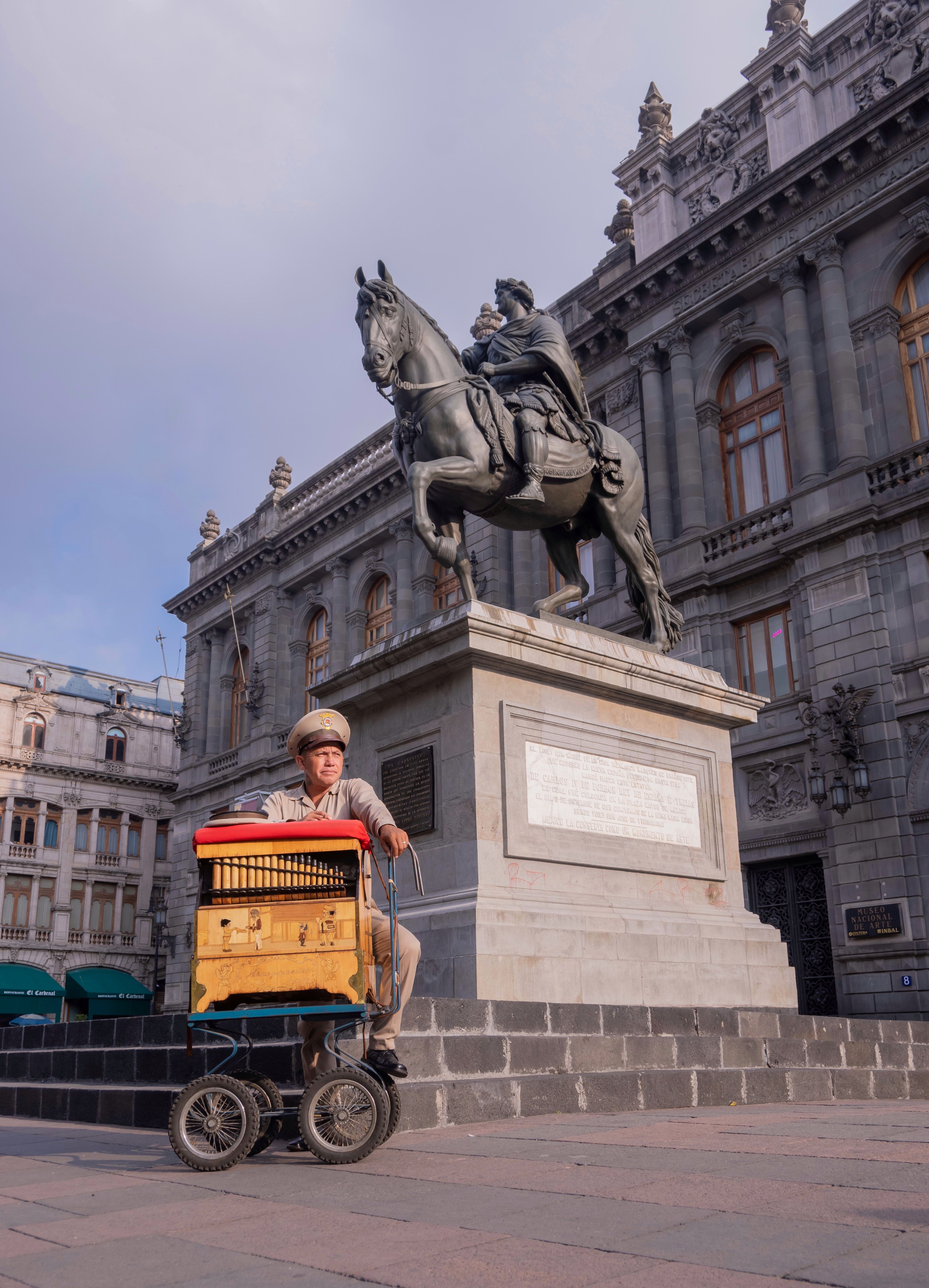 A traditional musician playing a barrel organ beside a grand equestrian statue in a historic city square.