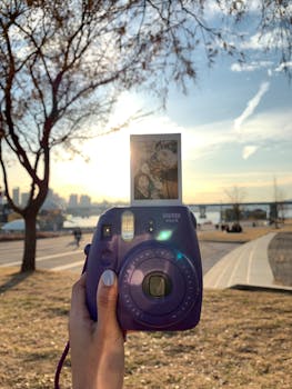 A purple Instax camera held against a scenic park sunset, capturing leisure moments.