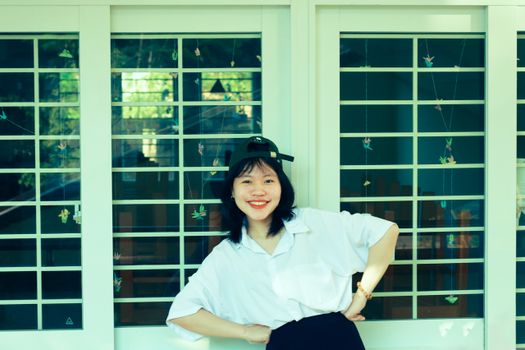 Smiling young woman with arms akimbo, posing in front of a window on a sunny day.