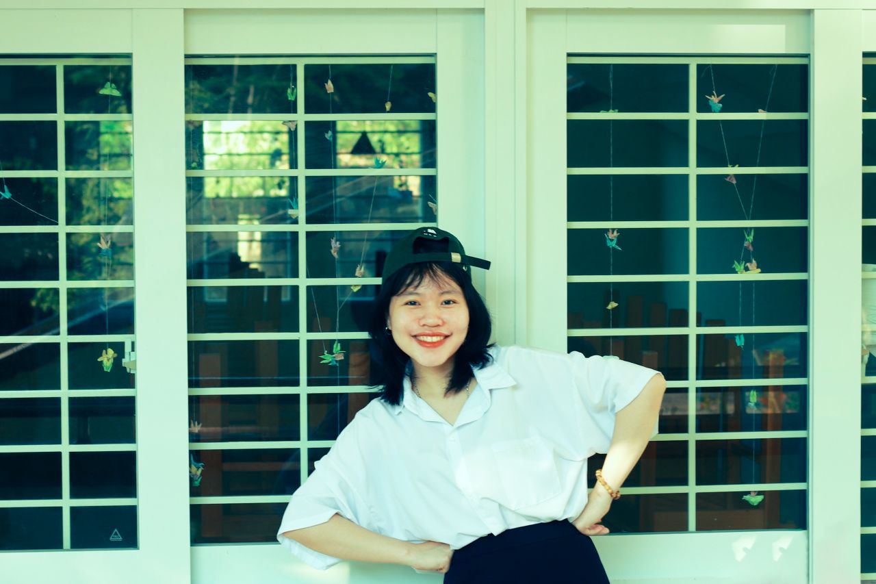 smiling young woman with arms akimbo posing in front of a window on a