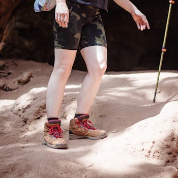 A hiker in camouflage shorts and boots walks on sandy terrain, using a trekking pole for support.