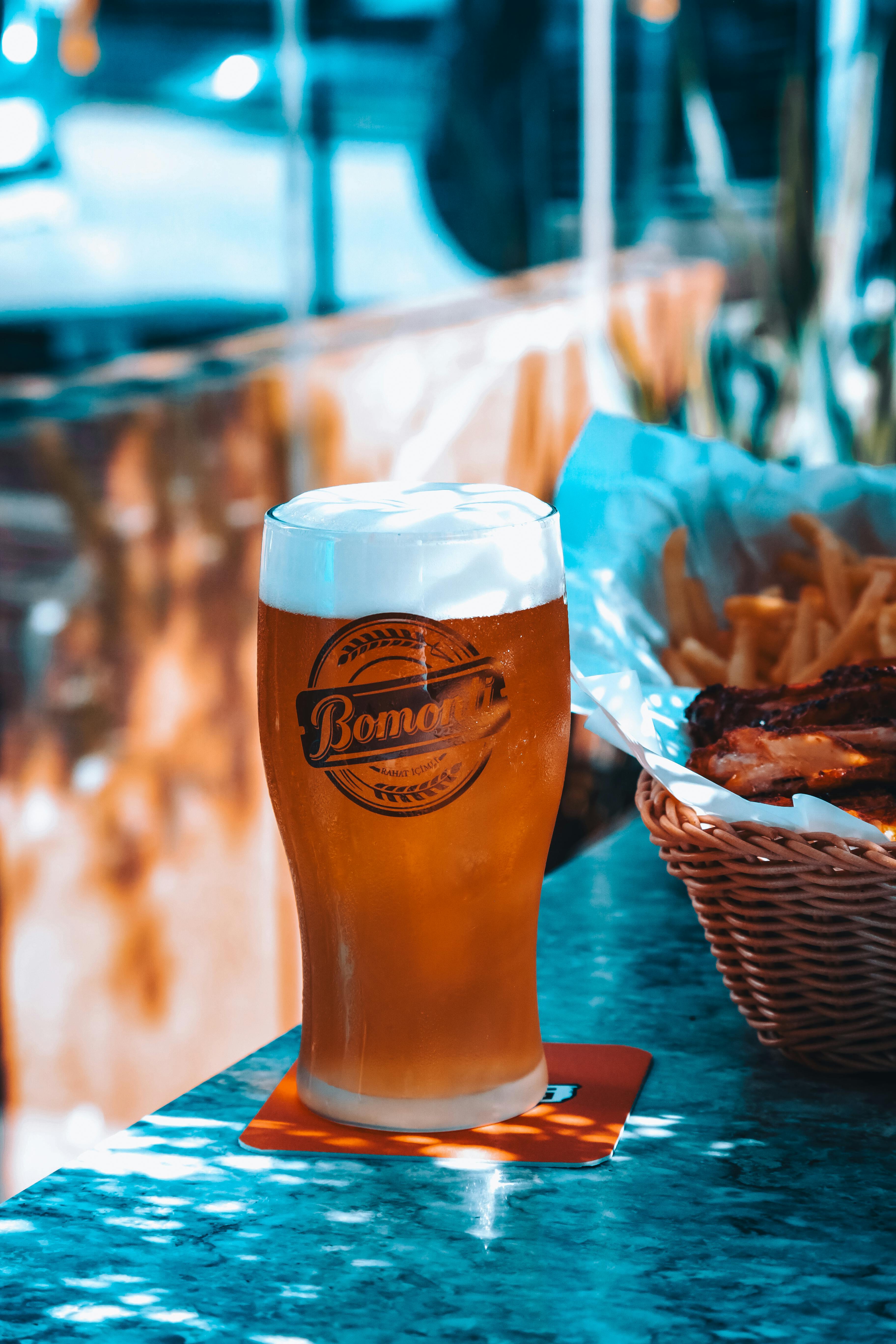 Close-up of a frothy beer glass on a sunny table with food in a basket.