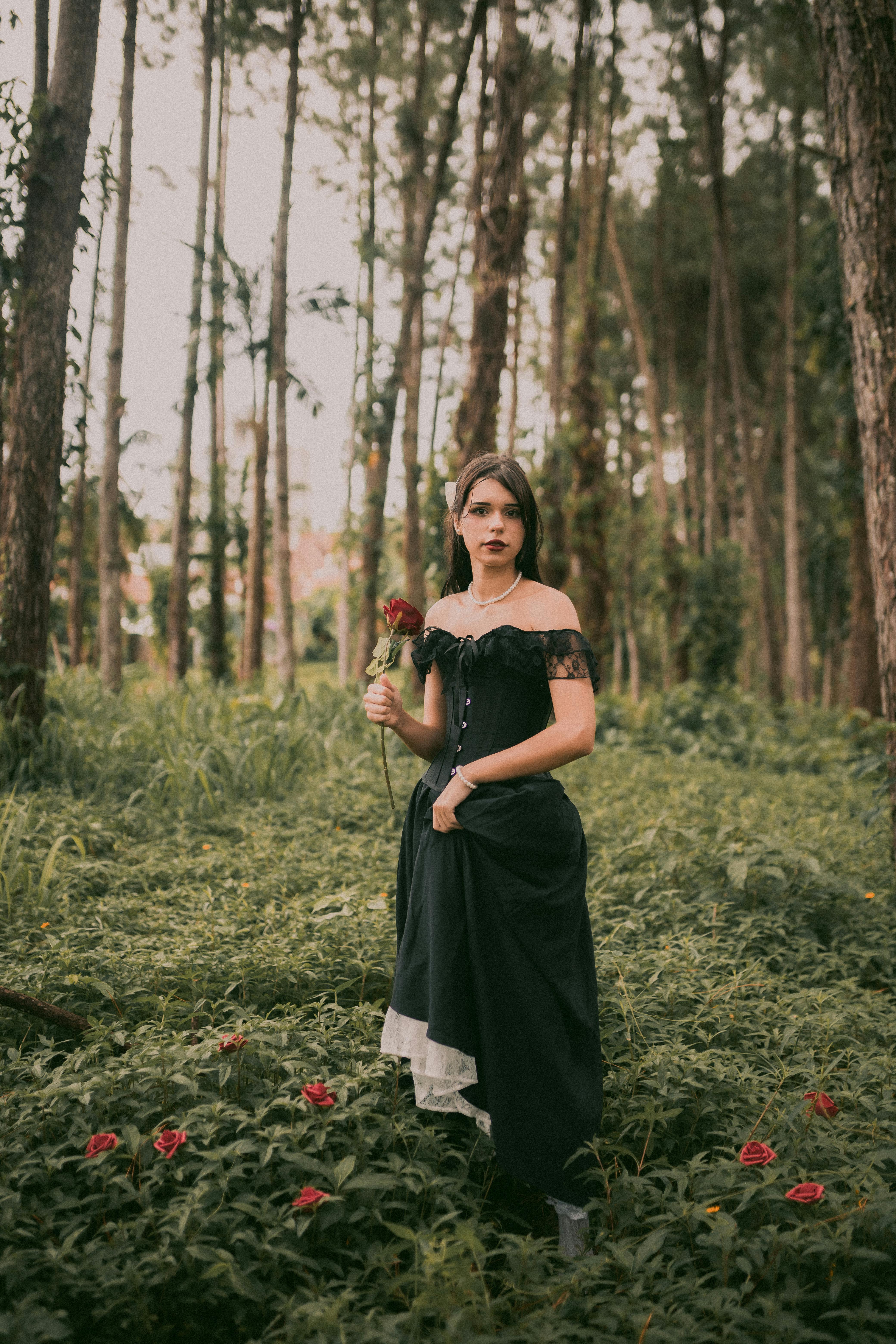 A young woman in a black dress holding a red rose amid a lush forest backdrop.