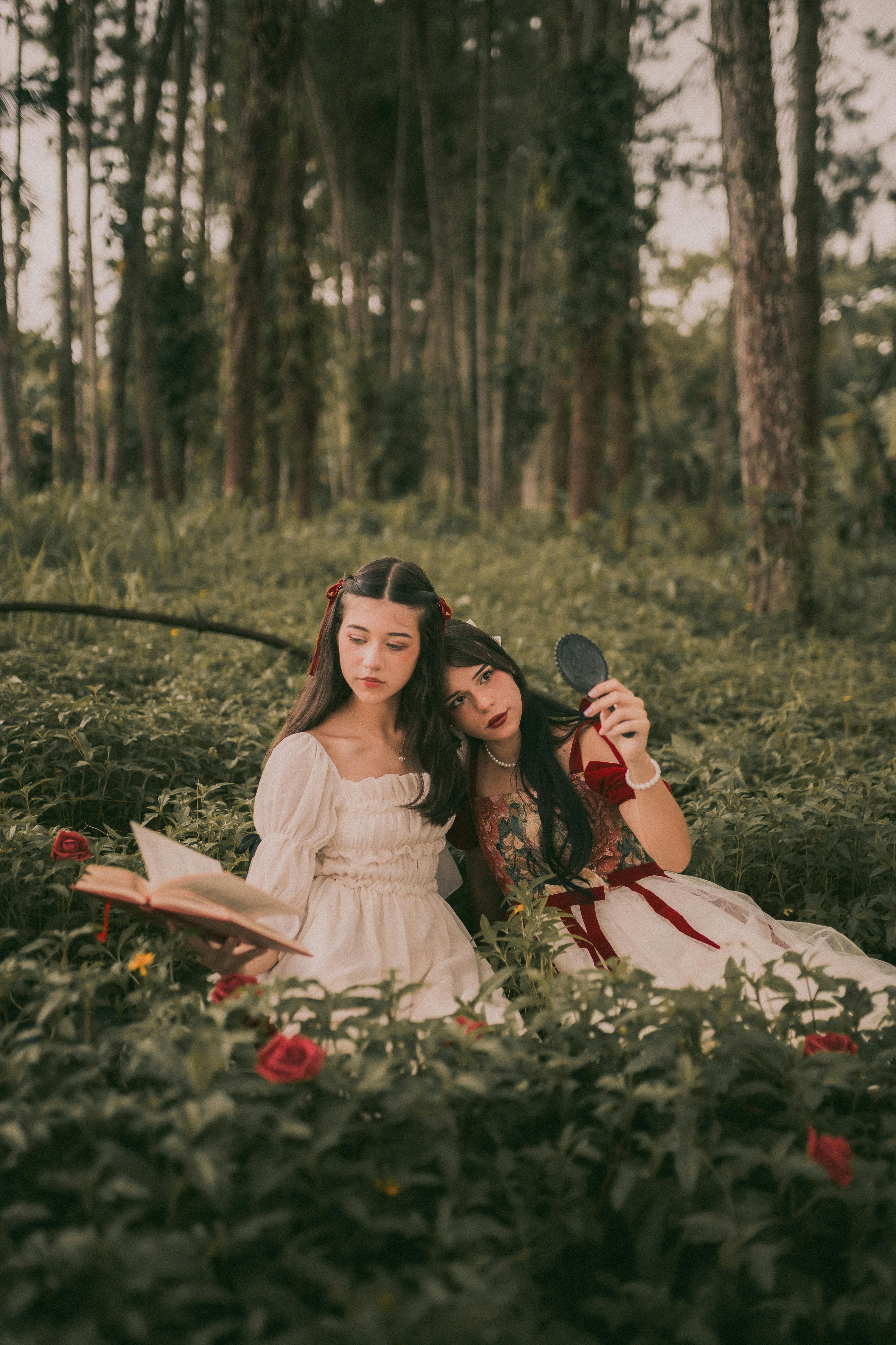 girls in dresses sitting on a meadow in a forest holding a book and a small mirror