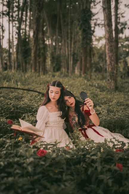 Two girls in fantasy dresses reading in a serene forest setting.
