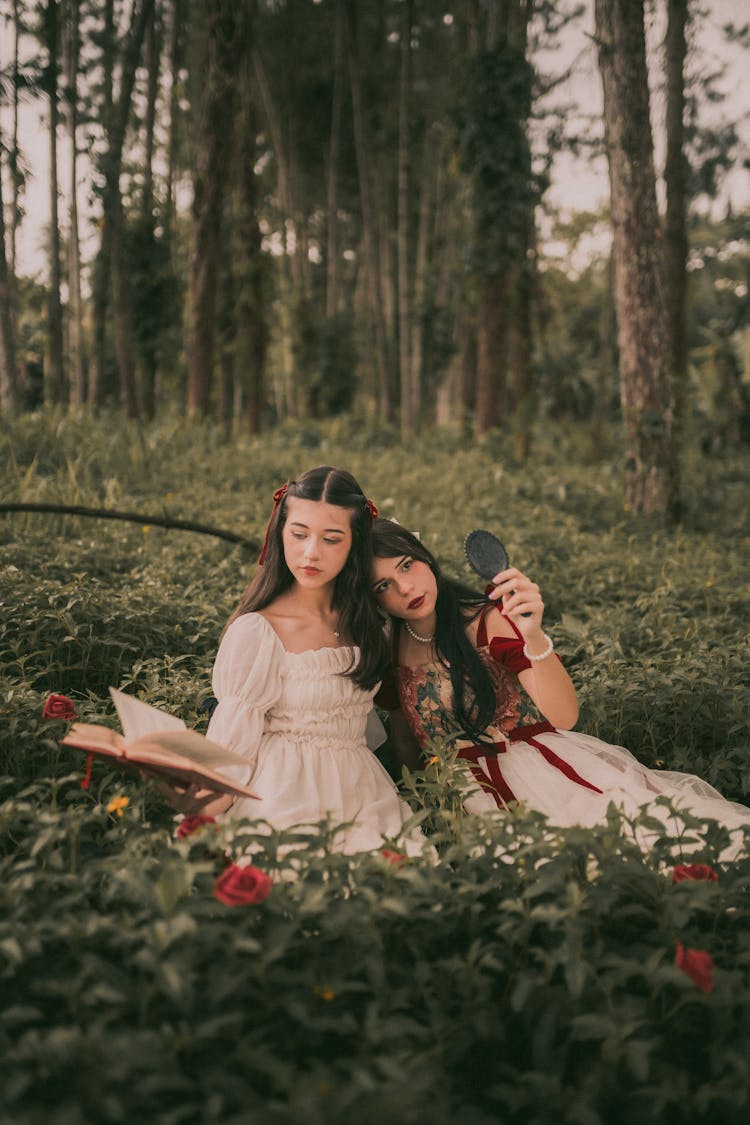 Girls In Dresses Sitting On A Meadow In A Forest, Holding A Book And A Small Mirror 