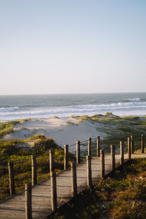 Free Serene view of a beach with wooden boardwalk, sand dunes, and waves crashing on the shore. Stock Photo