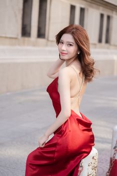 Charming Asian woman in a stylish red silk dress posing outdoors against urban architecture.