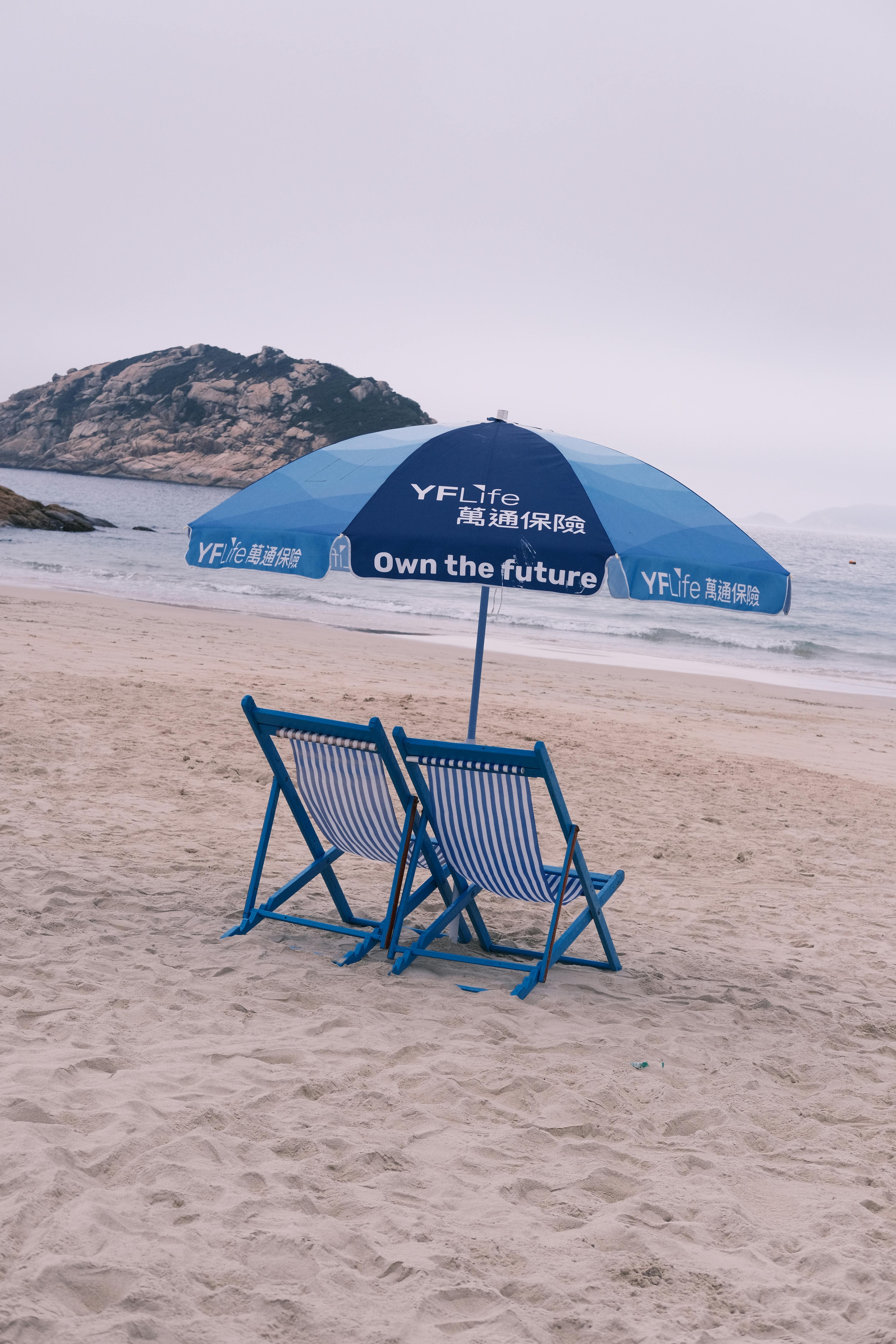 Peaceful beach setting with blue deckchairs under an umbrella facing the sea on a sandy shore.