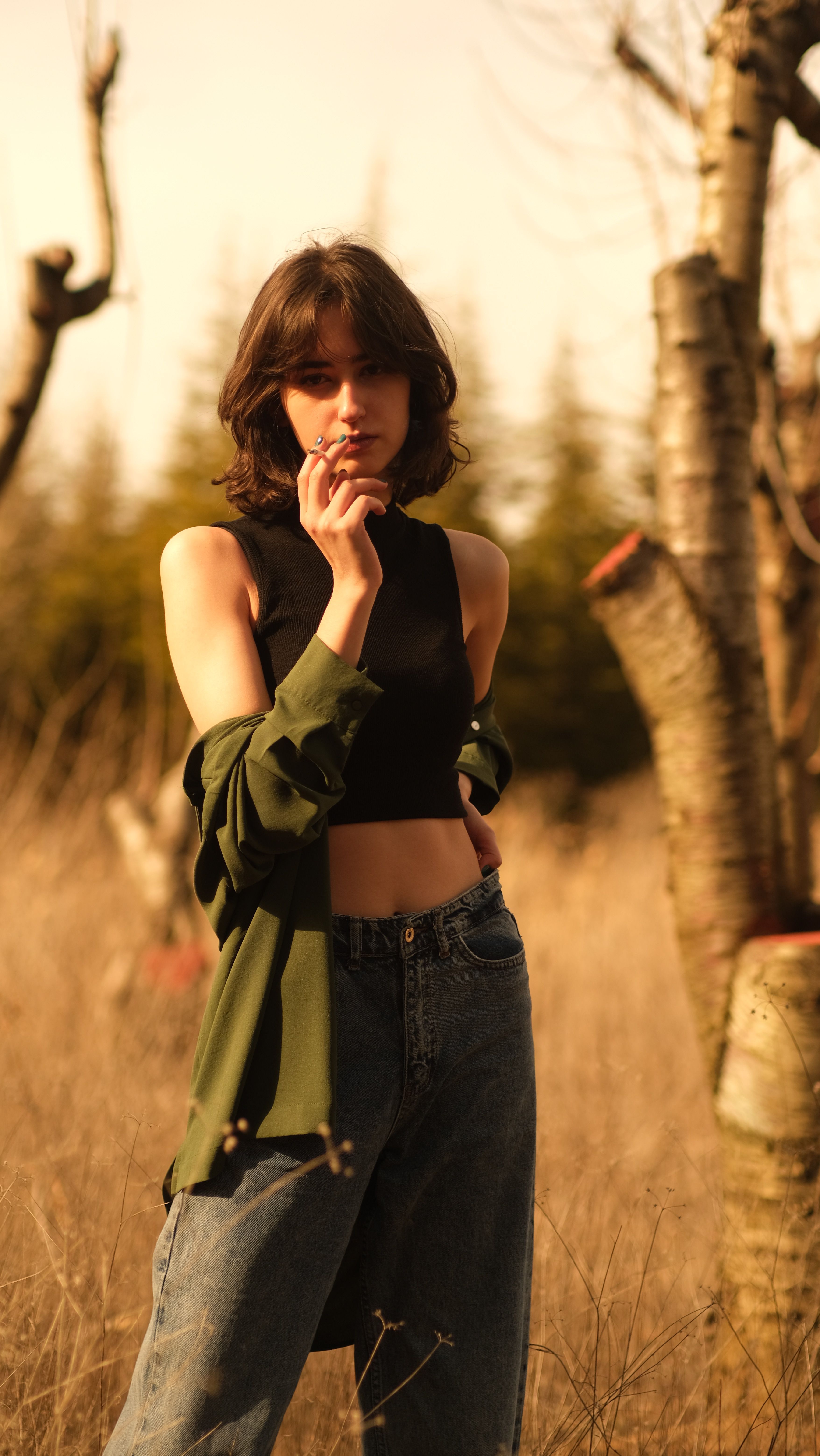 Young woman posing confidently in a sunlit meadow with trees, wearing casual attire.