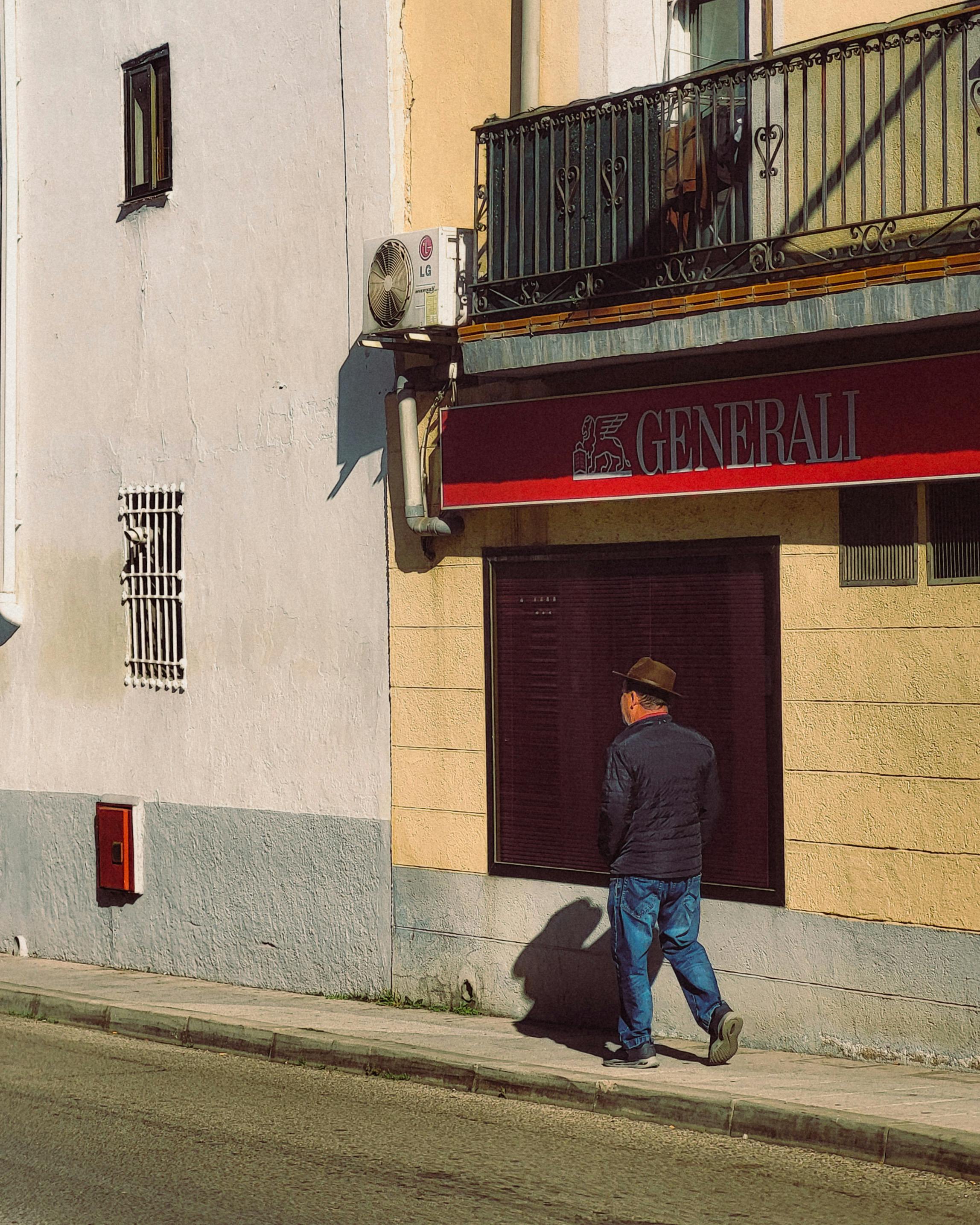 A man in a hat walks on a sunlit sidewalk next to a building with Generali signage.