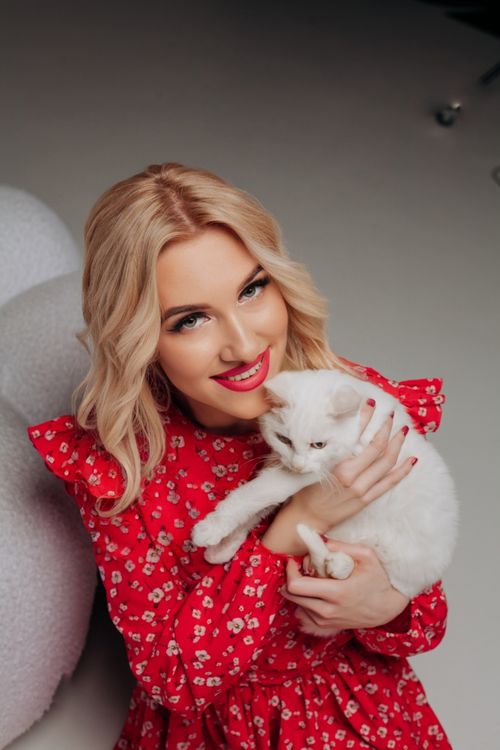 Free Smiling blonde woman in a red dress holds a white cat indoors. Stock Photo