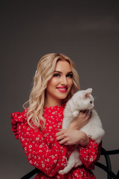Free Blonde woman in a red floral dress smiling while holding a white cat indoors. Stock Photo