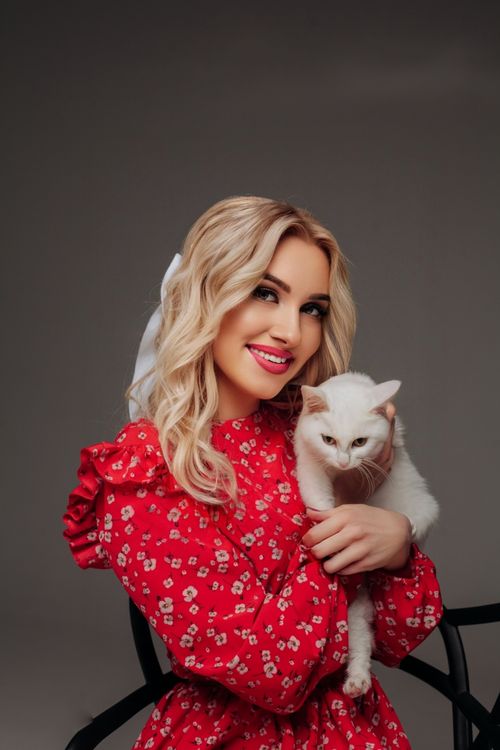 Free Blonde woman smiling in a red dress hugging a white cat against a simple background. Stock Photo