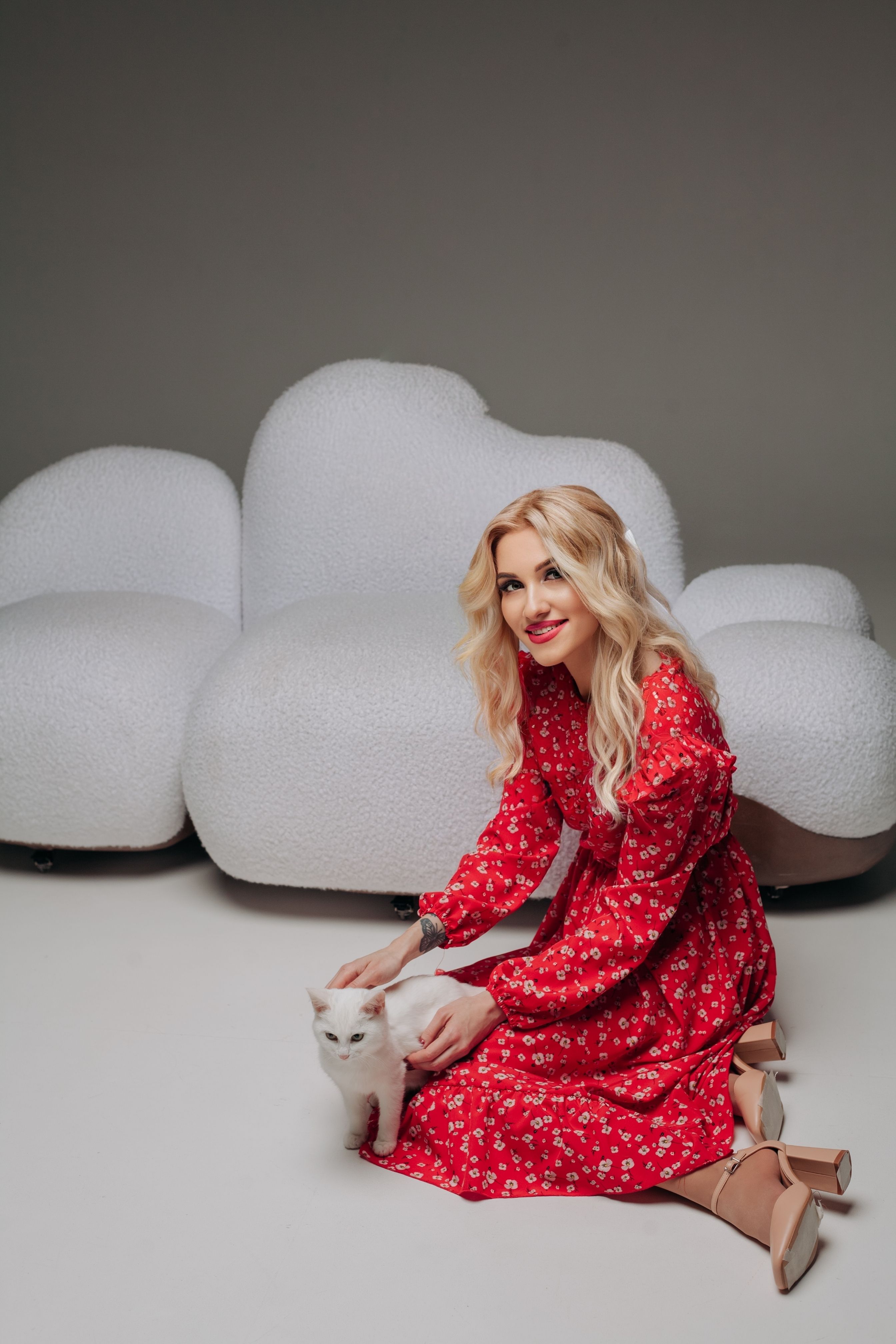 Blonde woman in a red dress posing with a white cat in an indoor studio setting.