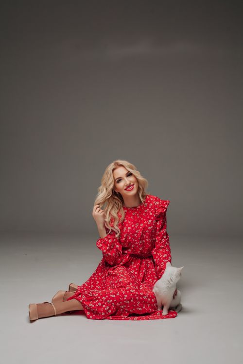 Free Blonde woman in red floral dress posing with white cat in a studio setting. Stock Photo
