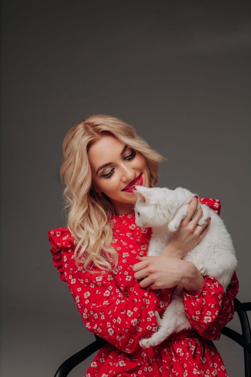 Free Smiling blonde woman in red floral dress holding a white cat in a studio. Stock Photo