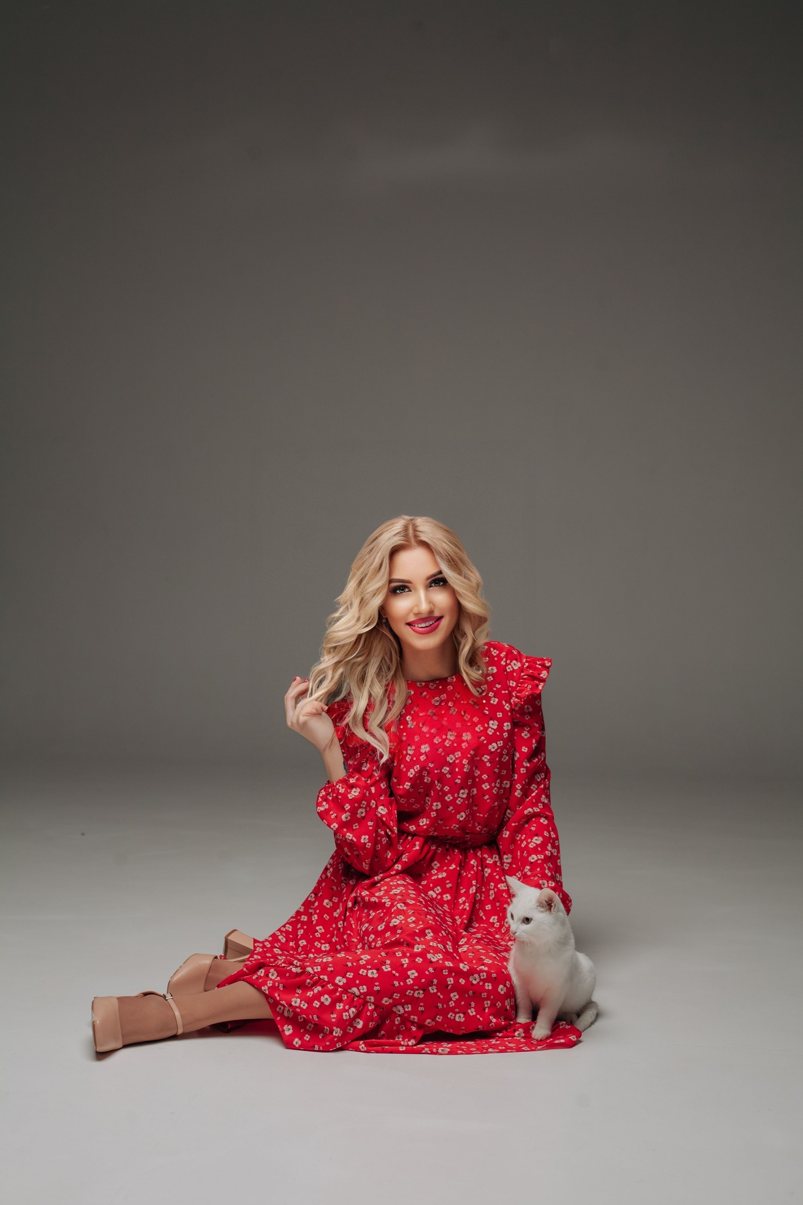 Stylish woman in red floral dress posing with a white cat in a studio setting.