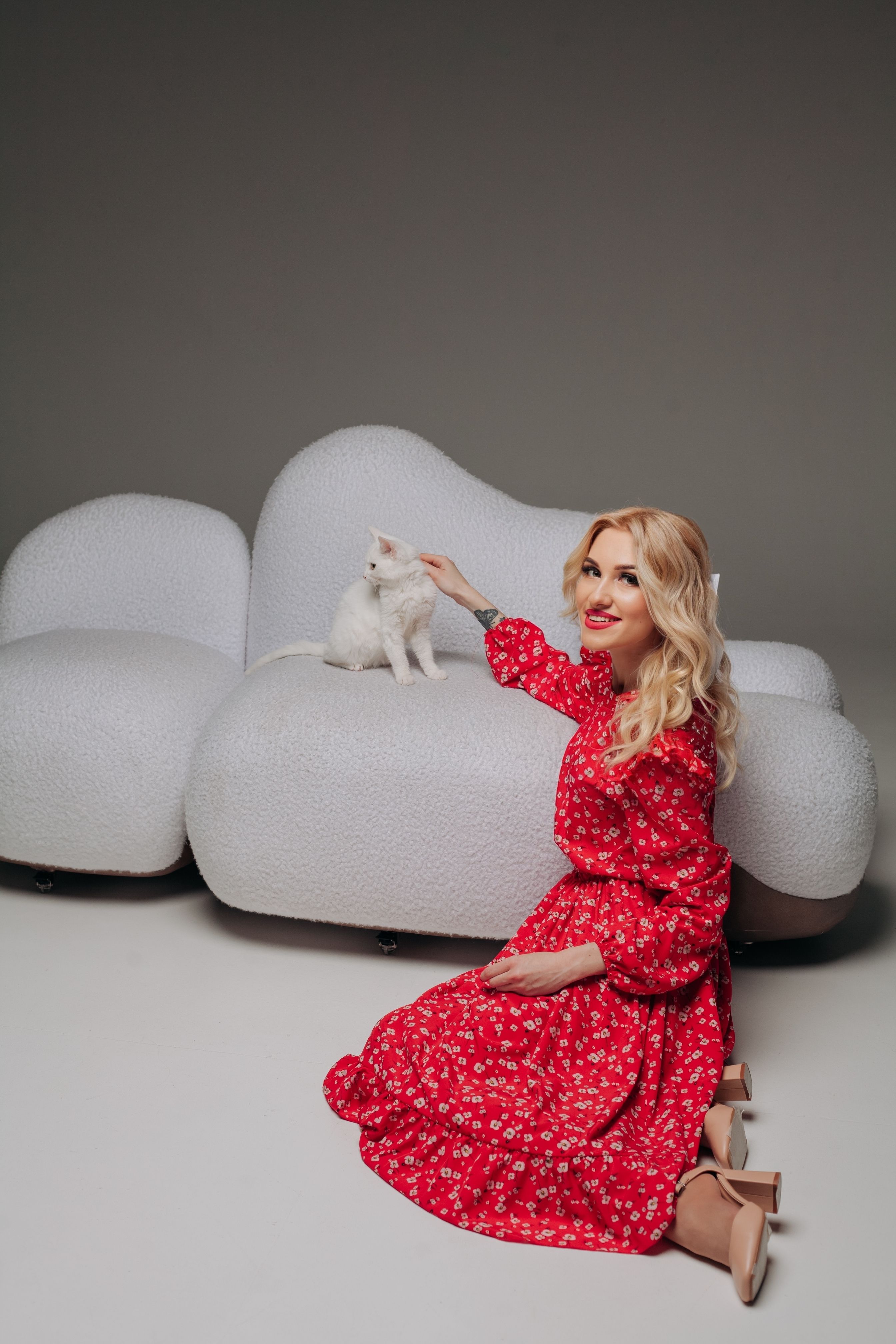 Stylish blonde model in red dress posing with white cat on a modern sofa in a studio setting.