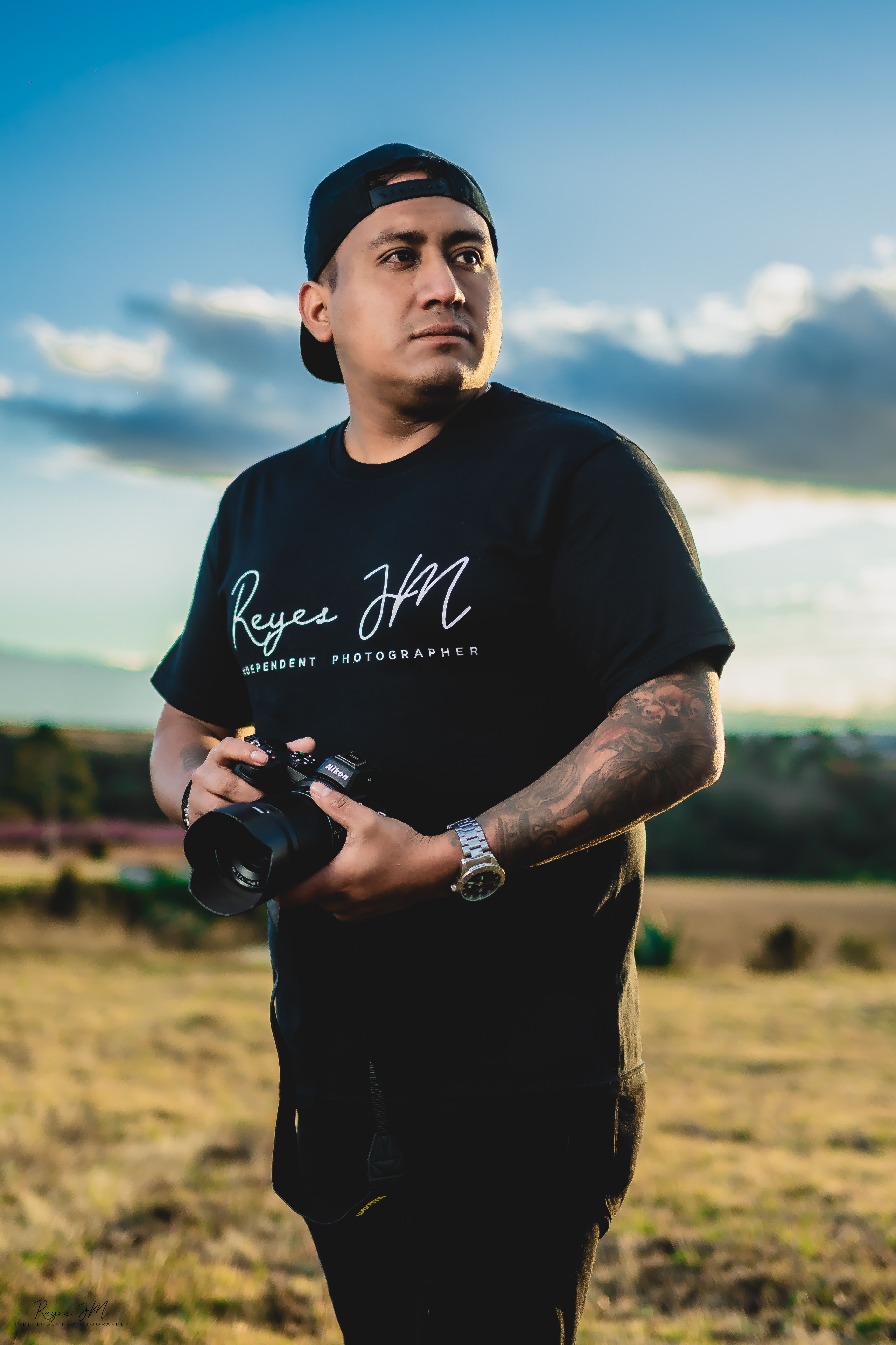 A man holding a camera stands in a grassy field with a cloudy sky backdrop.