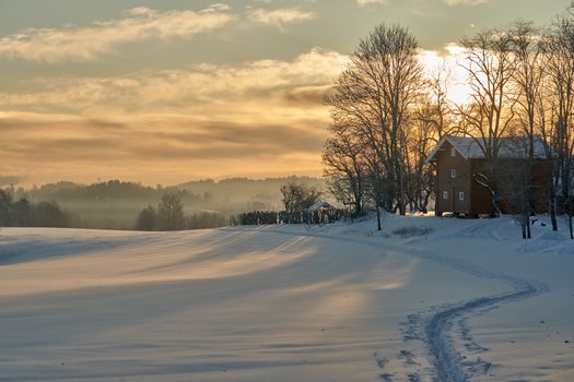 A tranquil scene of a snow-covered field with a sunset behind bare trees and a house.