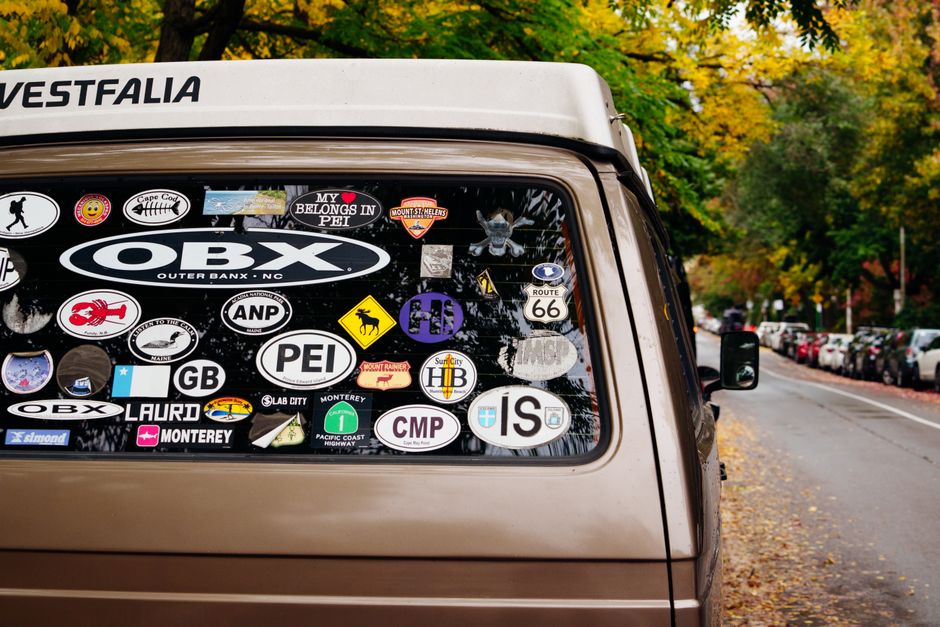 A vintage van adorned with various travel stickers parked on an autumn street with colorful foliage.
