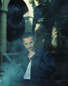 A man in a jacket stands thoughtfully amidst smoke, creating a moody atmosphere.