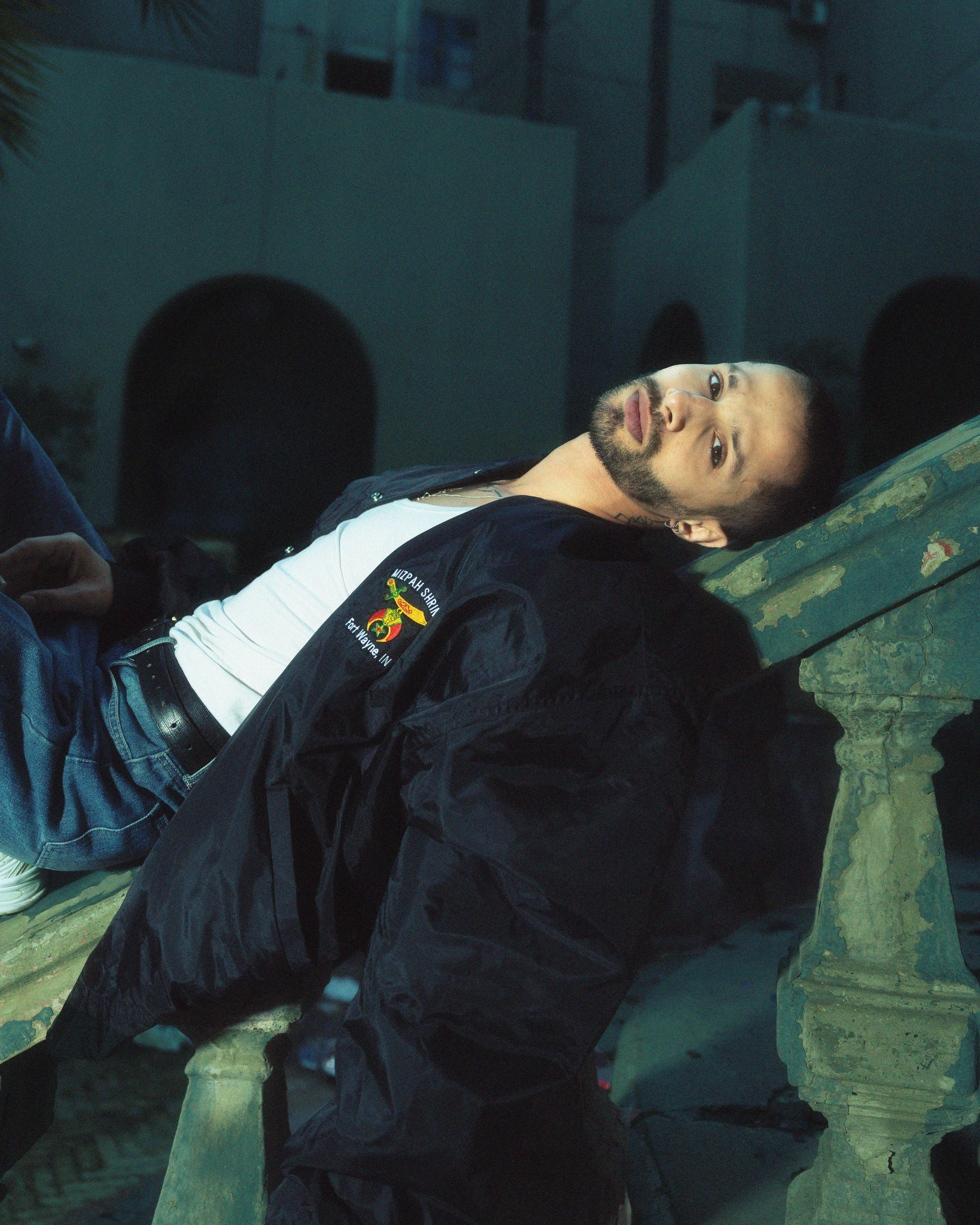 Man in a casual jacket reclining on an ornate railing, gazing upwards in a relaxed pose.