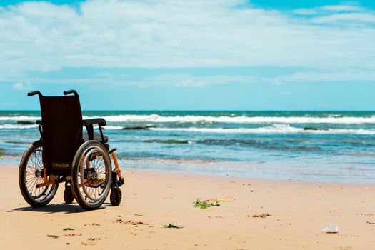 A wheelchair sits on a serene beach, facing the calming sea waves under a clear sky.