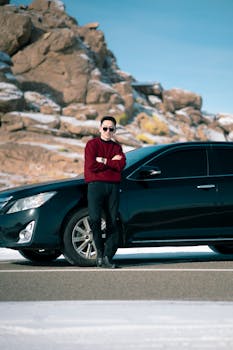 Explore the snowy mountain road with a stylish man standing beside a black car, showcasing travel and adventure.