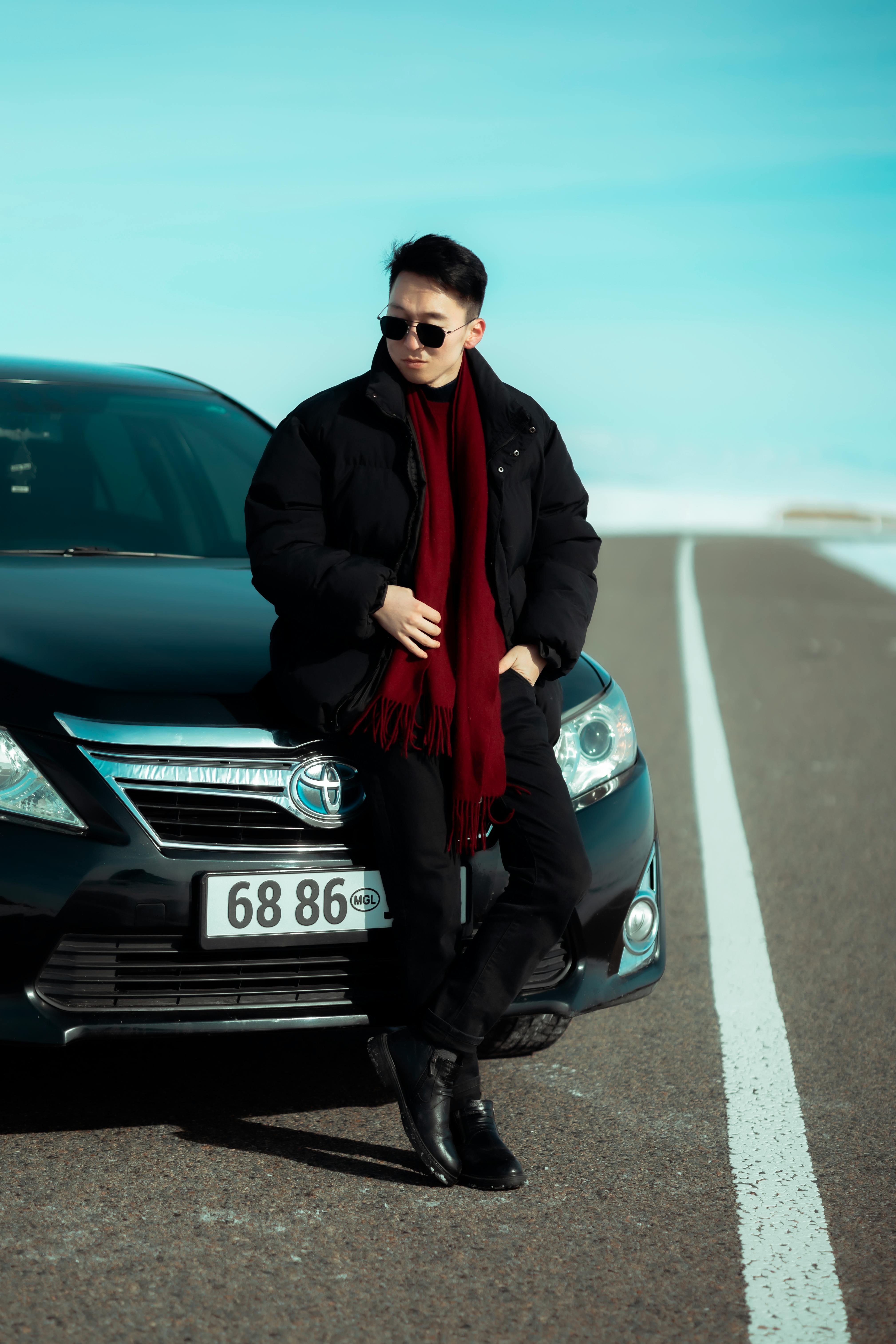 Young man wearing sunglasses and a red scarf leans on a luxury car on an open road.