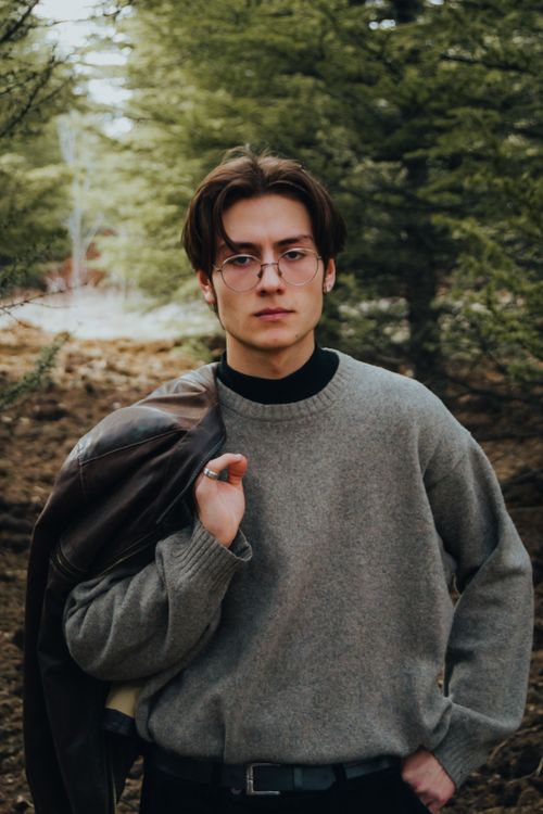 Free Portrait of a young man wearing glasses and a sweater, standing confidently in a forest setting. Stock Photo