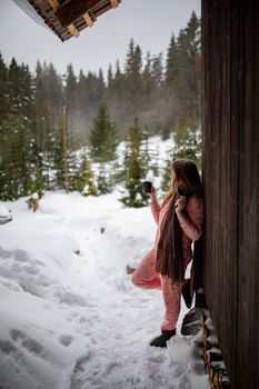 A woman enjoys coffee outdoors in pajamas, leaning against a snowy wooden cabin, surrounded by a forest in winter.