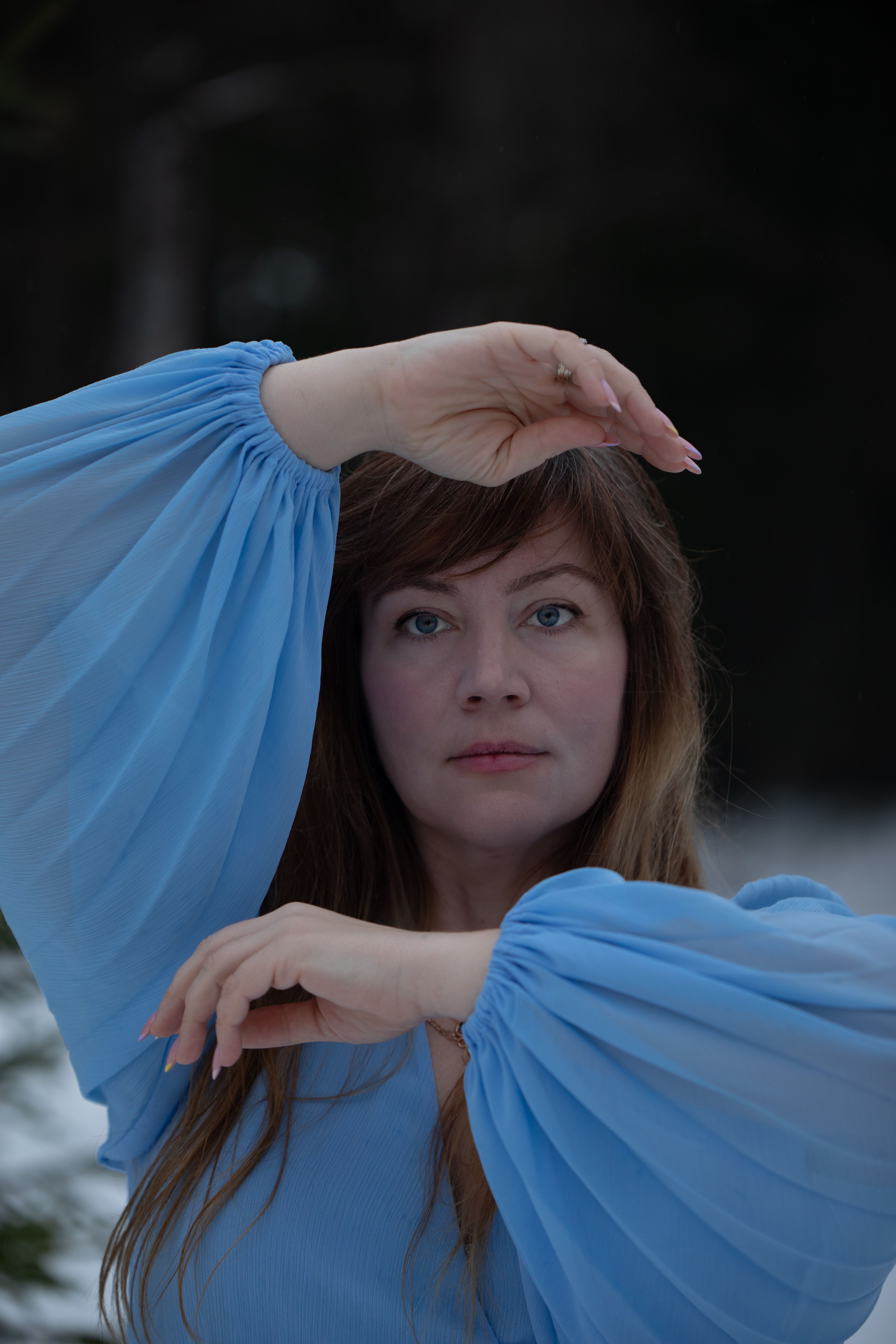 Portrait of a woman in a blue dress posing elegantly outdoors with hands around face.