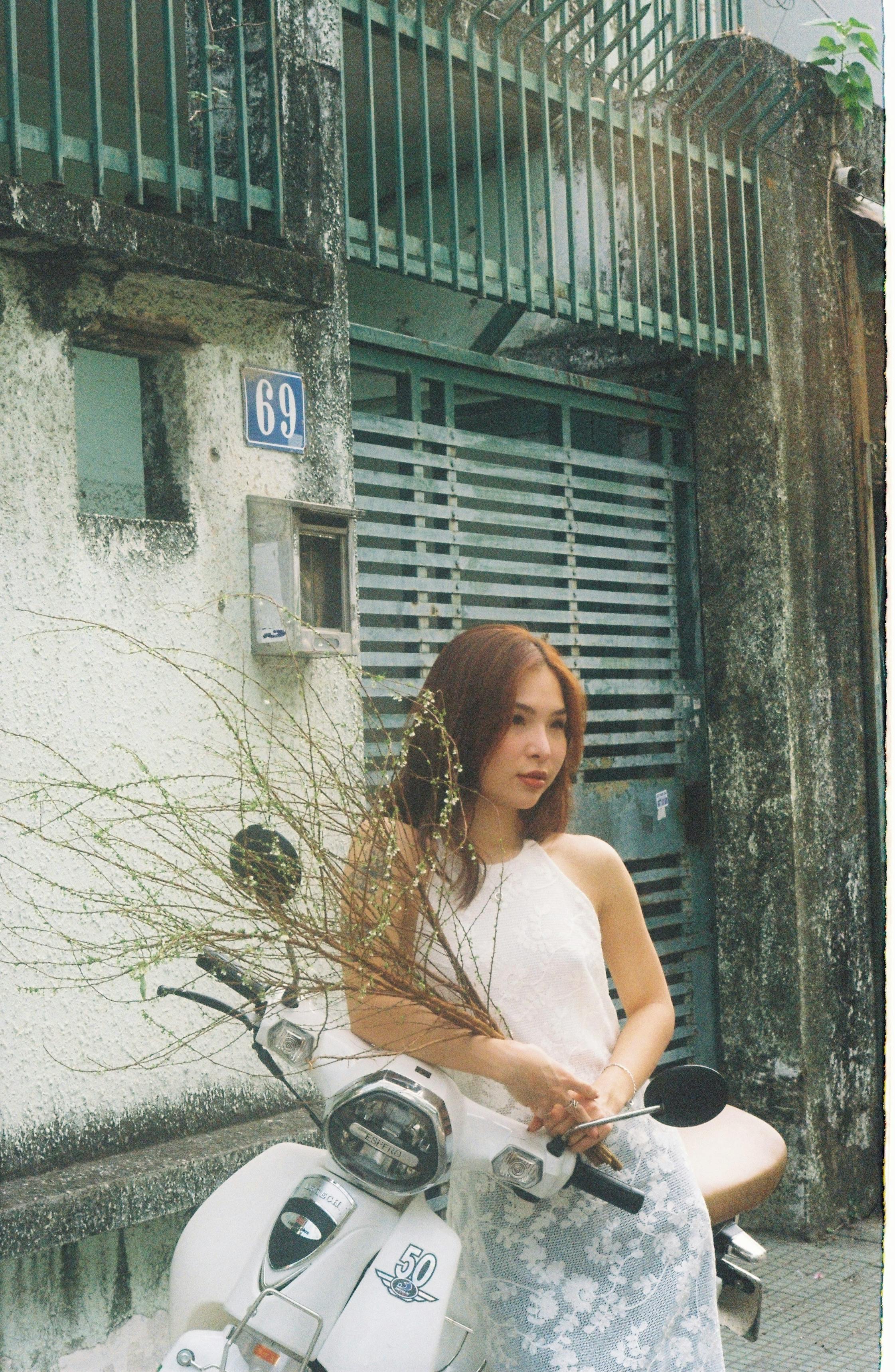 A young woman stands next to a vintage scooter and wall, holding twigs.
