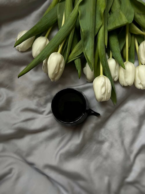 A black coffee cup and white tulips on a bed