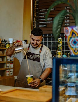 Barista serving freshly brewed coffee at café counter, wearing apron with menu board behind.