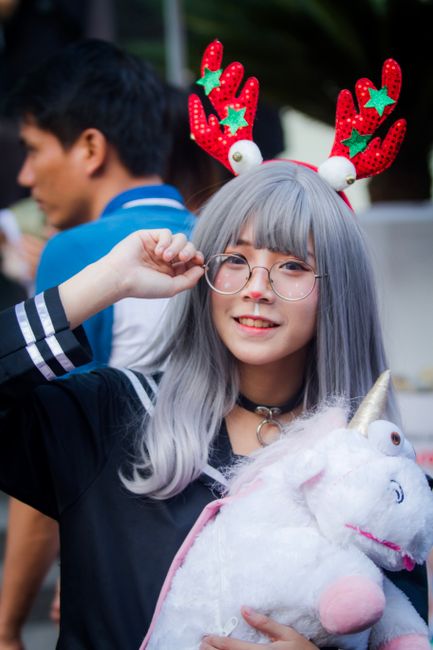 Young woman in cosplay with festive headdress and unicorn toy, smiling outdoors during the day.