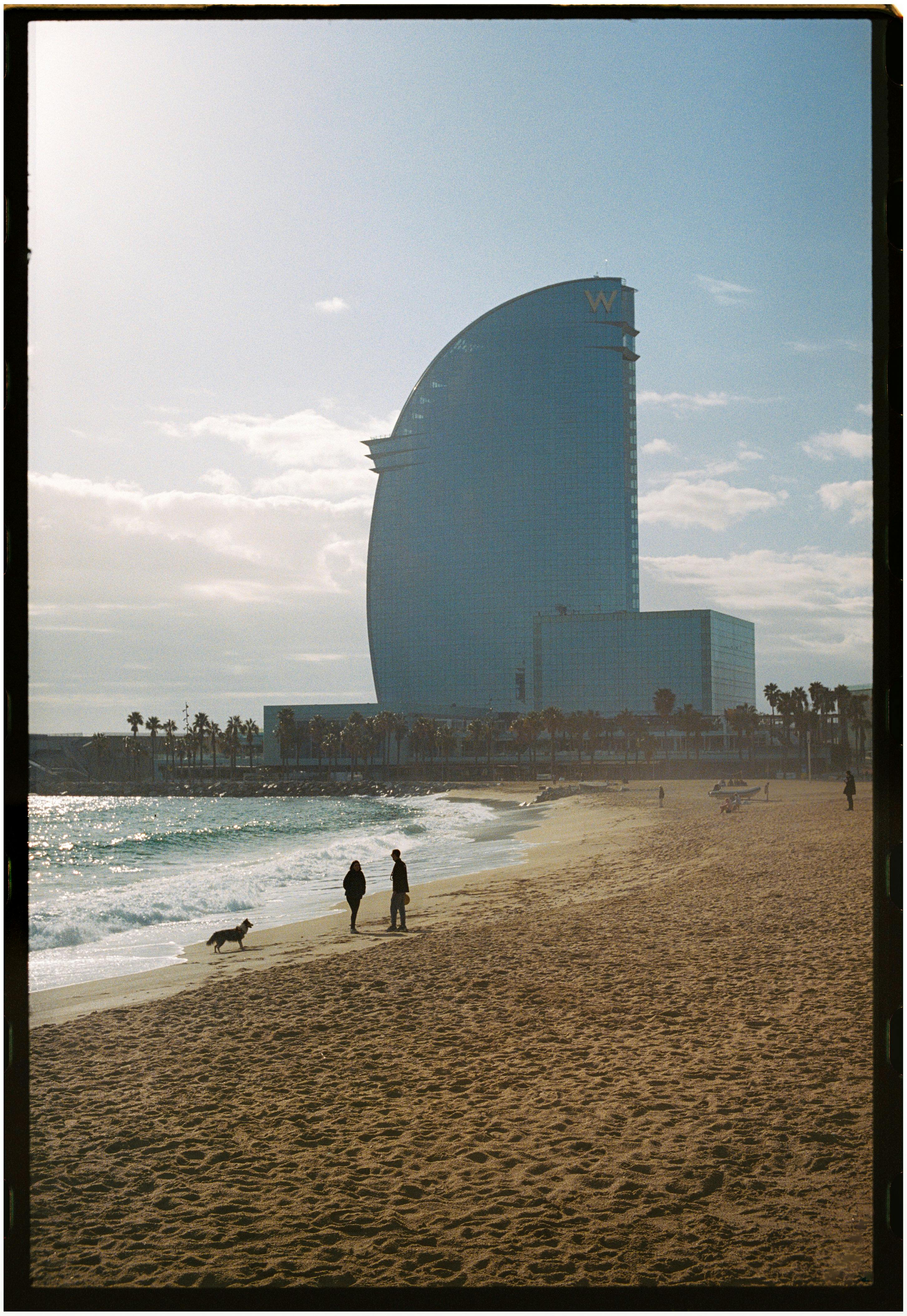 Two people and a dog on a beach by the iconic W Barcelona, a remarkable architectural landmark.