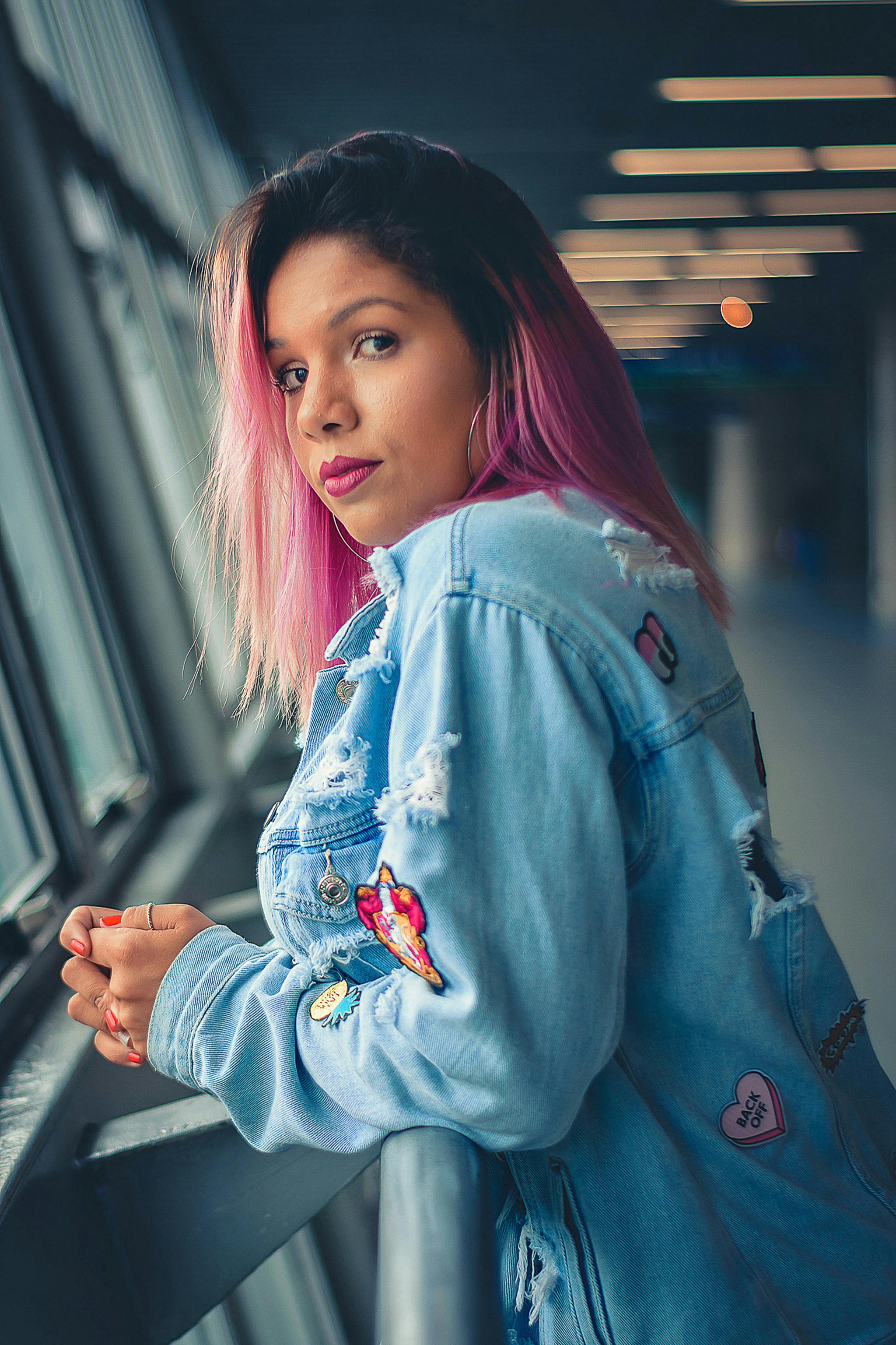 一名粉发时尚女子身穿牛仔夹克，靠在室内金属栏杆上摆姿势