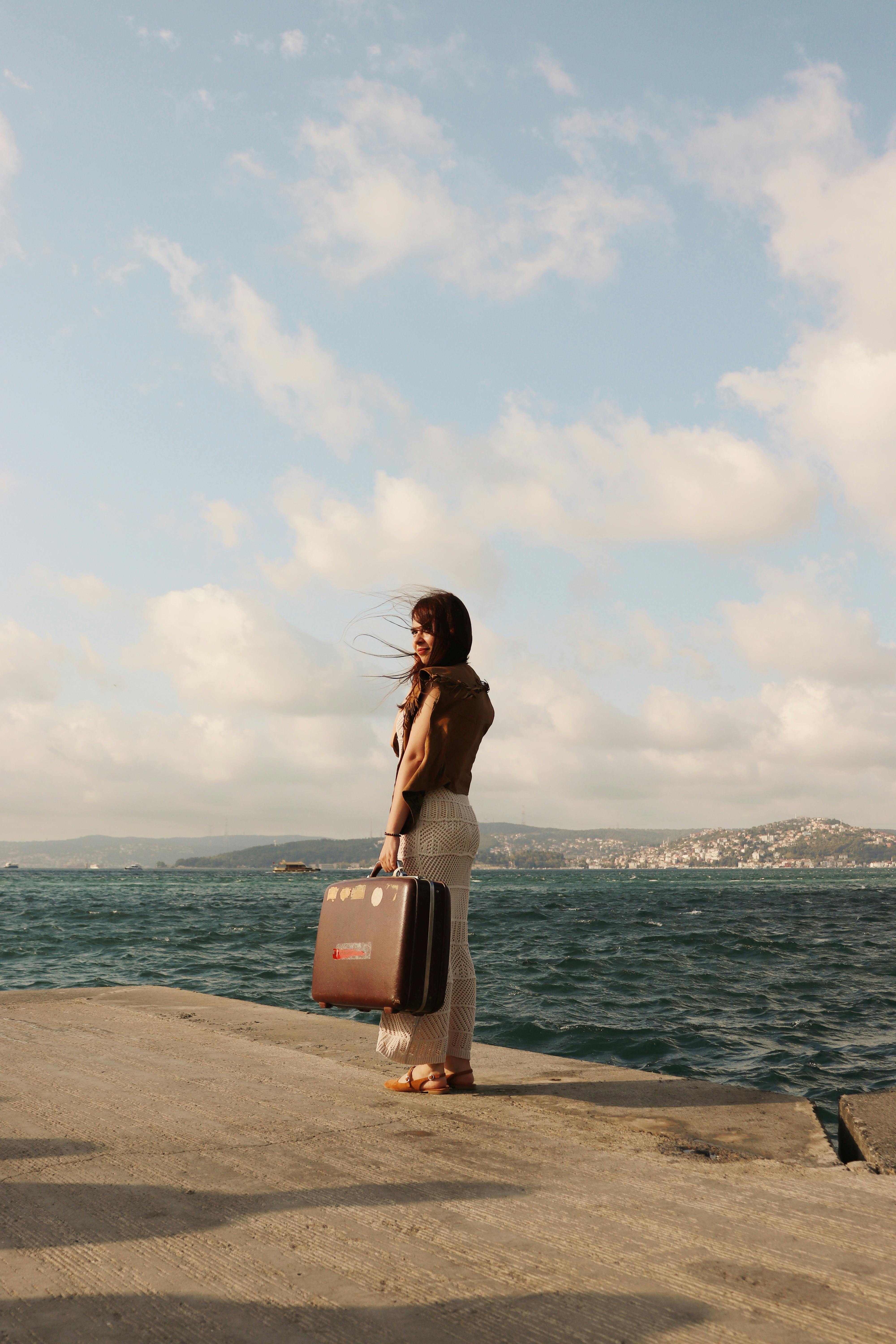 A woman stands with a suitcase on a pier by the sea, under a partly cloudy sky.