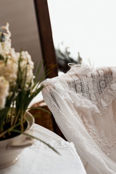 Delicate white lace dress draped over a chair with soft floral decor in a calm, warm setting.