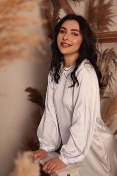 Smiling woman in white satin dress posing elegantly inside a bohemian setting.