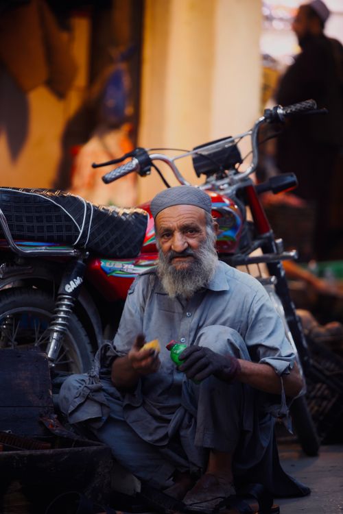 Free Elderly man with a beard sits by a motorcycle on a city street, holding an object in a vibrant urban scene. Stock Photo